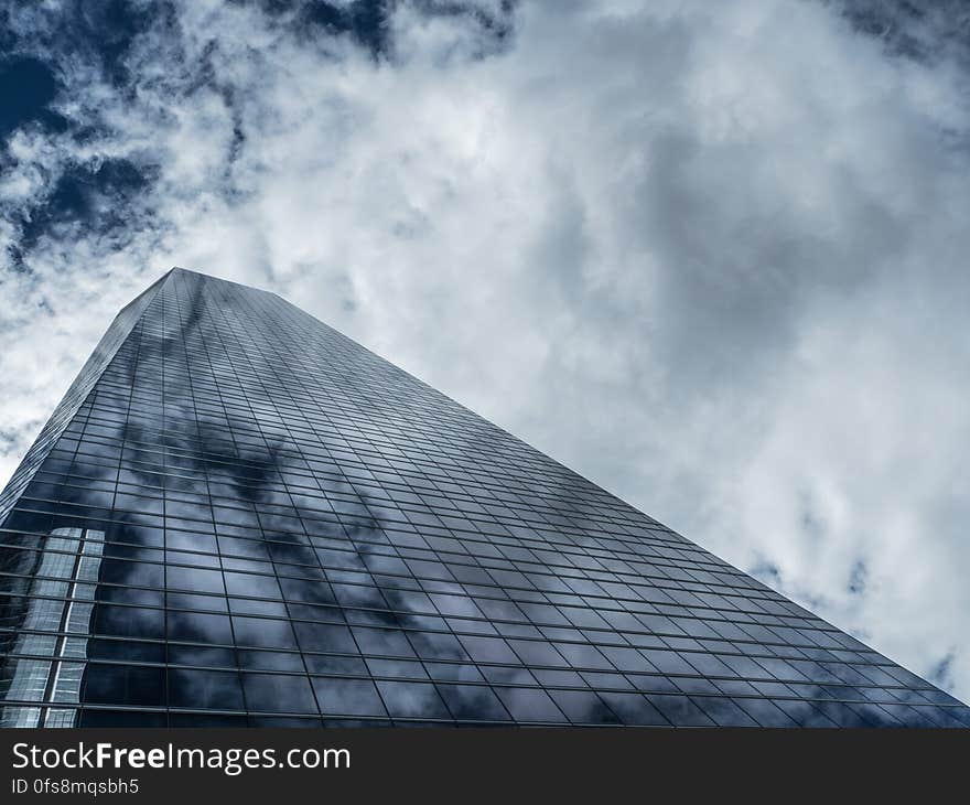 View pointing upwards towards the top of a skyscraper, constructed of mainly steel and glass with light filtering through clouds casting a shadowy pattern on the facade of the building. View pointing upwards towards the top of a skyscraper, constructed of mainly steel and glass with light filtering through clouds casting a shadowy pattern on the facade of the building.