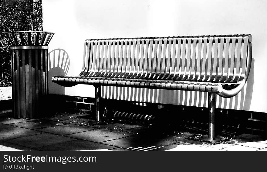 A black and white image of a bench and a trashcan by a wall. A black and white image of a bench and a trashcan by a wall.