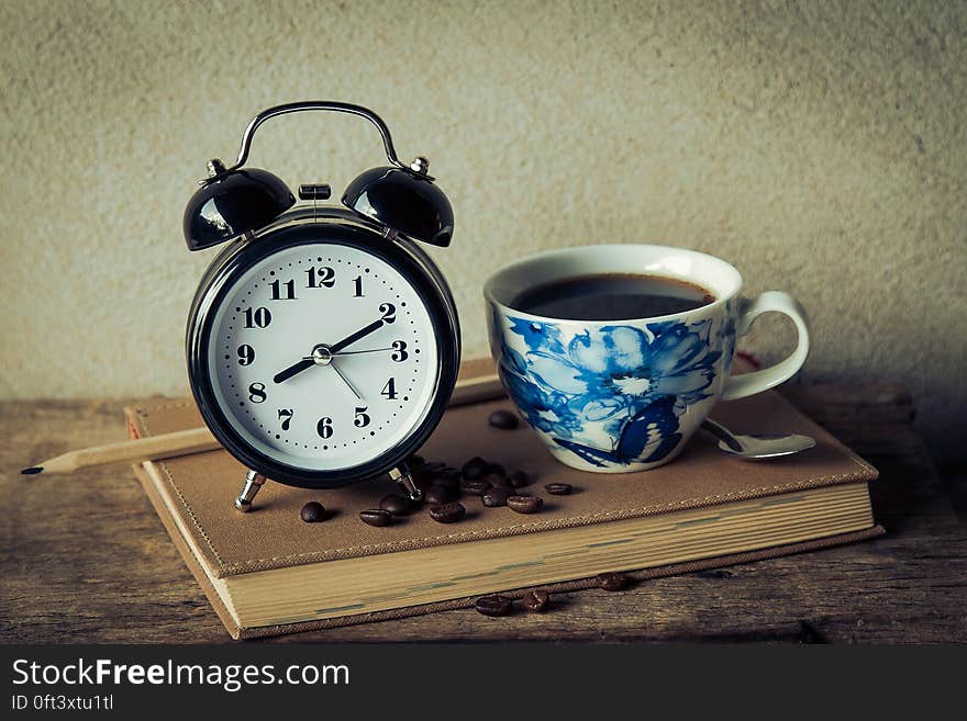 Still life of vintage alarm clock on leather book with pencil and coffee cup on rustic wooden tabletop. Still life of vintage alarm clock on leather book with pencil and coffee cup on rustic wooden tabletop.