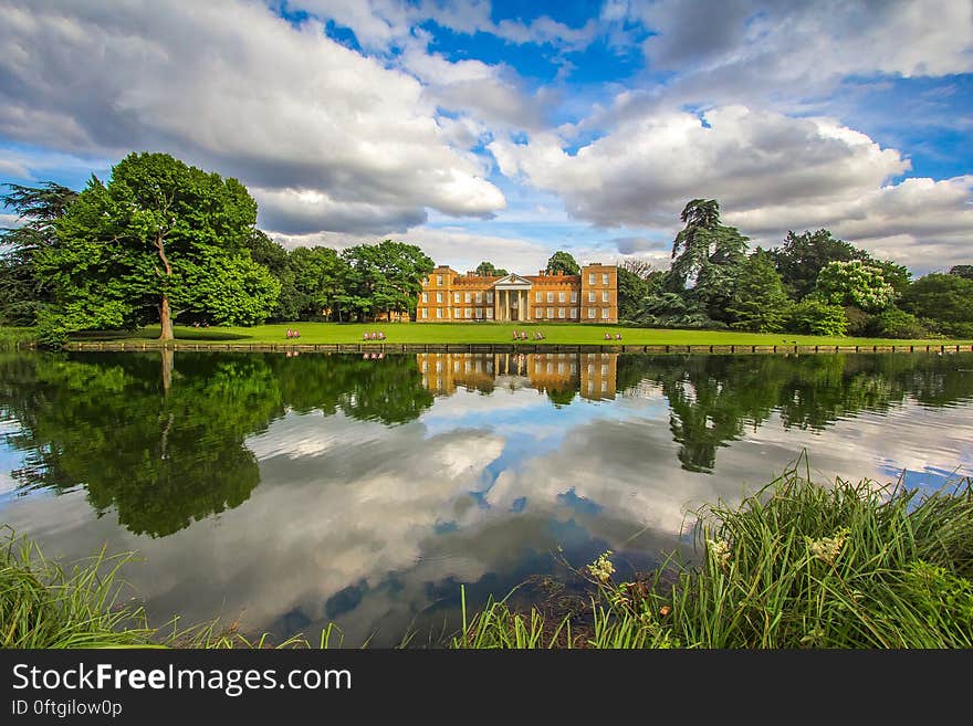 Brown and White House on Green Grass Lawn Surrounded by Trees in Front of Lake Under Blue and White Cloudy Skyt