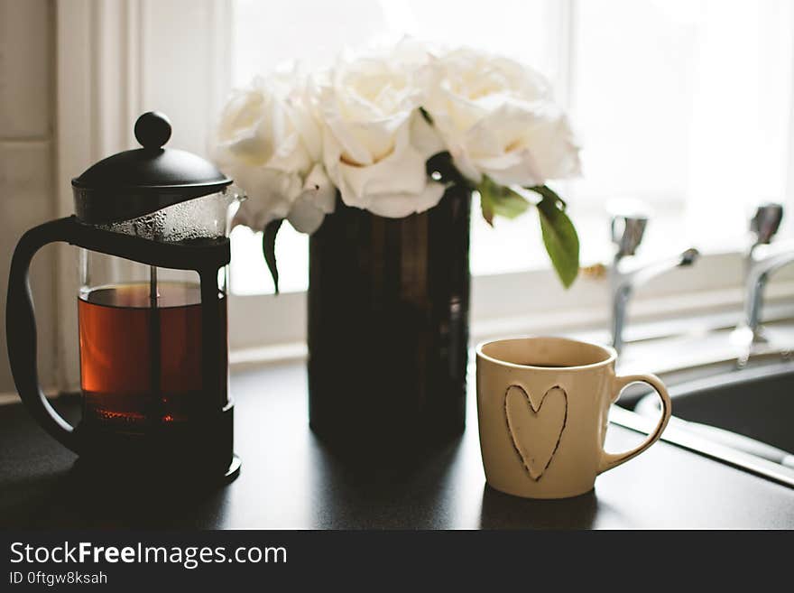 A French press coffee or tea maker and cup with flowers on a kitchen table.