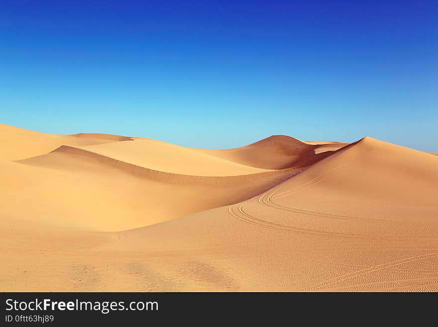 Sand dunes in the desert with vehicle tracks possibly indicating a tourist adventure or safari, cloudless blue sky.