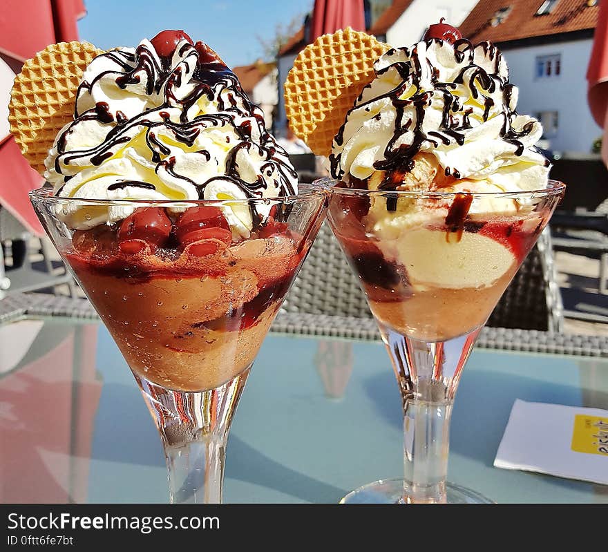 A pair of cocktail glasses with ice cream, cherries, whipped cream and sauce on a restaurant table.