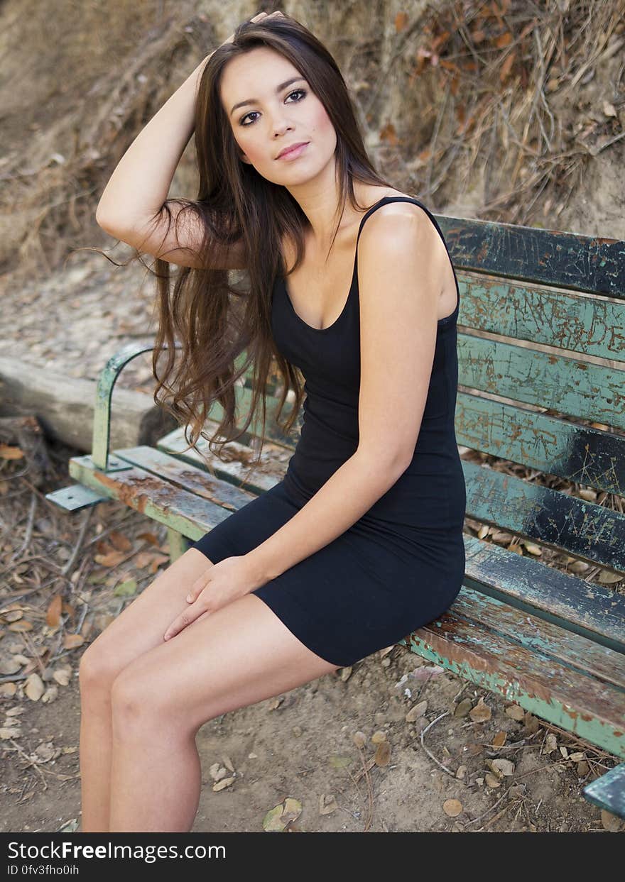 Portrait of young woman with long black hair wearing black dress sitting outdoors on park bench. Portrait of young woman with long black hair wearing black dress sitting outdoors on park bench.