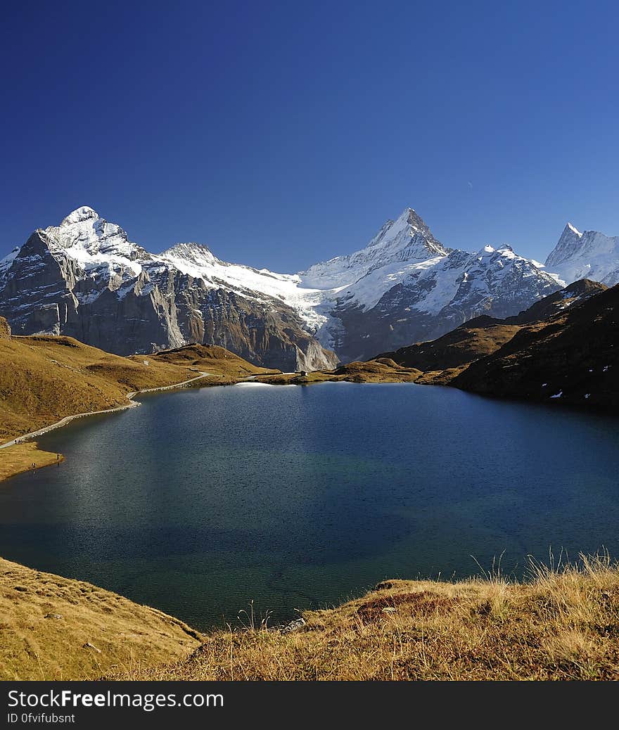 Blue waters of a sunlit lake with several snow covered mountain peaks beyond it, cloudless blue sky. Blue waters of a sunlit lake with several snow covered mountain peaks beyond it, cloudless blue sky.