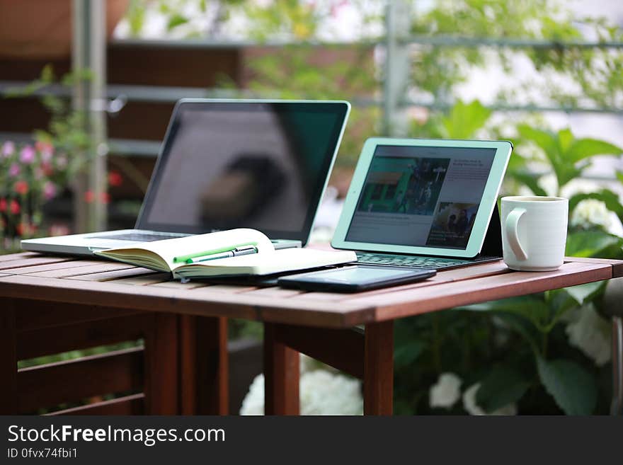 Laptop computers and coffee mug with open notebook on wooden table in sunny garden. Laptop computers and coffee mug with open notebook on wooden table in sunny garden.