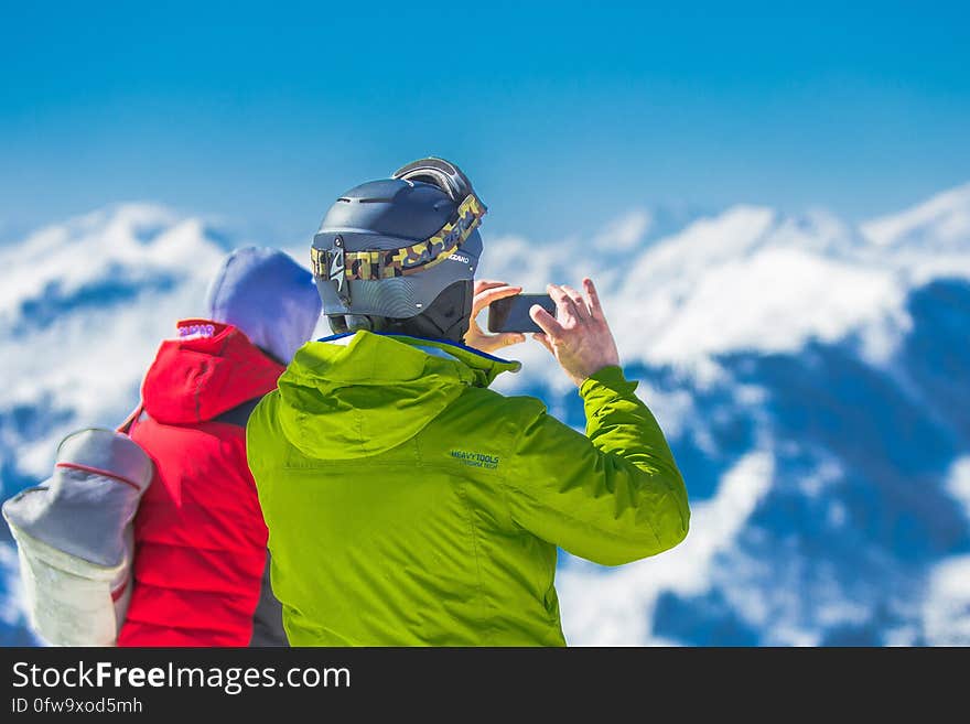 Man in Green Jacket and Gray Helmet Holding Phone Standing Next to Person in Red and Black Jacket