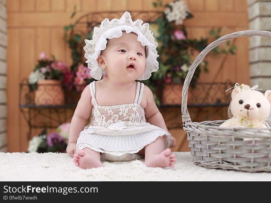 A young baby sitting on table wearing a white dress and a hat. A young baby sitting on table wearing a white dress and a hat.