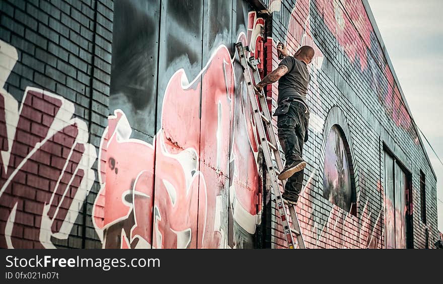 Man in Gray Shirt Standing on Gray Steel Ladder Painting Black White and Red Graffiti on Concrete Wall Outdoors