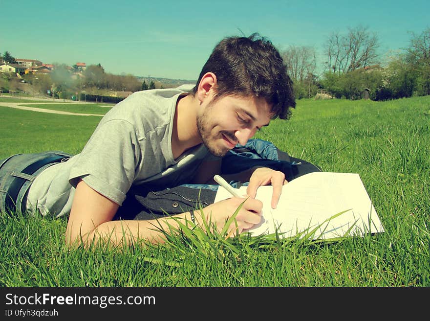 Young dark haired man lying almost prone in a green grassy field but writing (or drawing) on a sheet of white paper, blue sky. Young dark haired man lying almost prone in a green grassy field but writing (or drawing) on a sheet of white paper, blue sky.