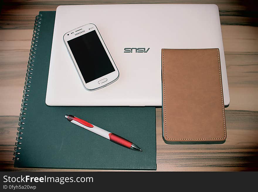 Spiral notebook, laptop computer, mobile phone and leather case and red and white pen on a wooden table. Spiral notebook, laptop computer, mobile phone and leather case and red and white pen on a wooden table.