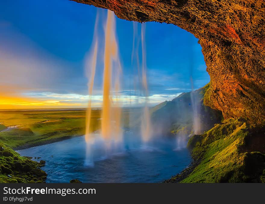 View, in golden light of a sunset, from a cave with water cascading down in front of the cave into a small pool, blue sky. View, in golden light of a sunset, from a cave with water cascading down in front of the cave into a small pool, blue sky.