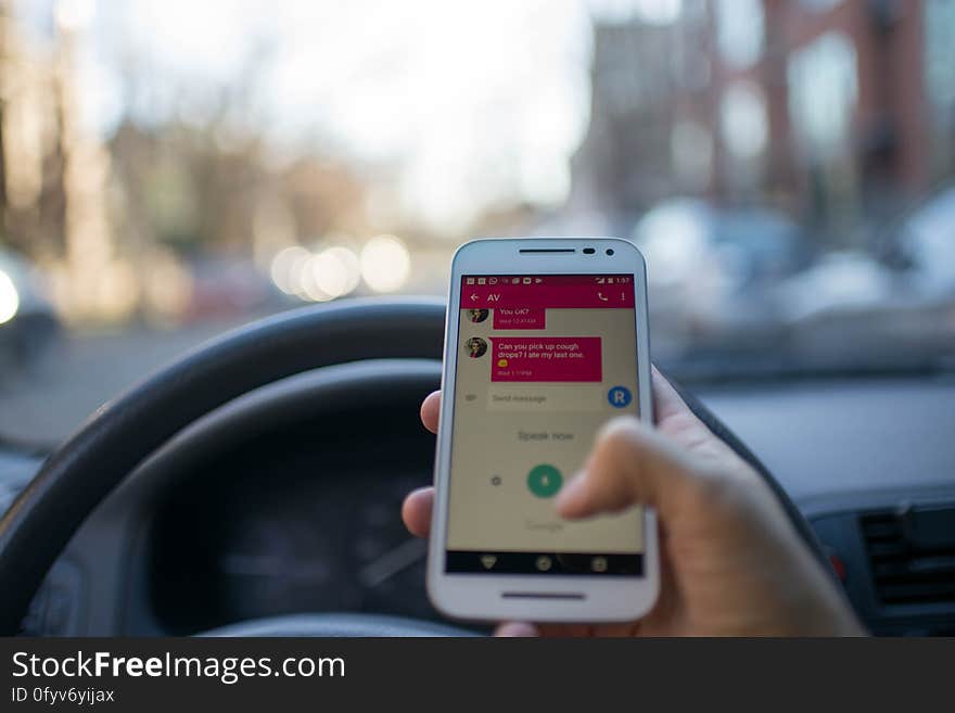 A person behind the steering wheel of a car with a smartphone in hand. A person behind the steering wheel of a car with a smartphone in hand.