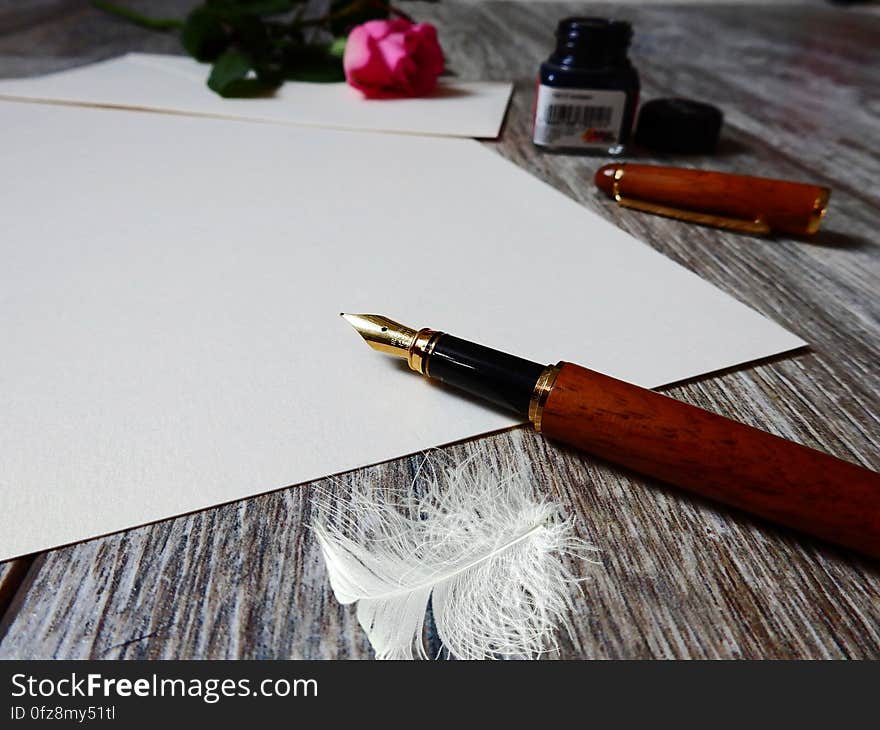 Fountain pen on blank sheet of paper with jar of ink, rose and feather on rustic wooden tabletop. Fountain pen on blank sheet of paper with jar of ink, rose and feather on rustic wooden tabletop.