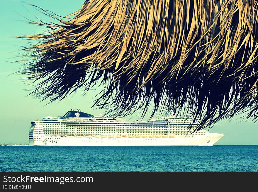 View under thatched beach umbrella of cruise ship in blue waters on sunny day. View under thatched beach umbrella of cruise ship in blue waters on sunny day.