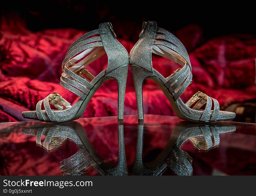 Side view of pair of silver shoes with stiletto heels reflecting on surface with red material in background.