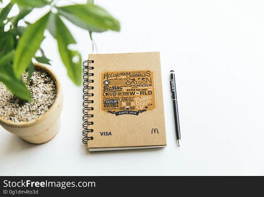 A notebook with a pen on a table with a houseplant. A notebook with a pen on a table with a houseplant.