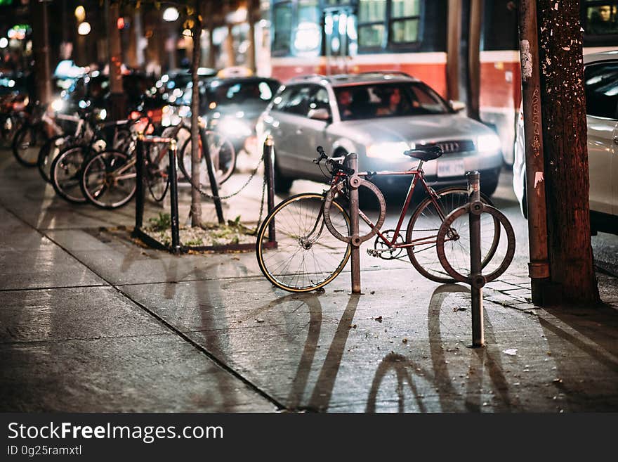 Bicycles parked at the side of a street with cars passing close by, their headlights casting long shadows over the wet sidewalk. Bicycles parked at the side of a street with cars passing close by, their headlights casting long shadows over the wet sidewalk.