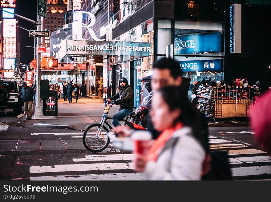 Urban road with cyclist riding over a pedestrian crossing with shops and shoppers in the background and a blurred anonymous couple on the pavement or sidewalk closeup. Urban road with cyclist riding over a pedestrian crossing with shops and shoppers in the background and a blurred anonymous couple on the pavement or sidewalk closeup.