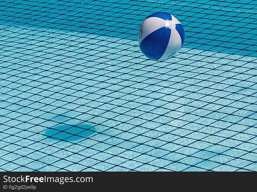 A blue and white beach ball floating on the surface of a swimming pool with tiled bottom. A shadow of the ball is cast on the bottom of the pool. A blue and white beach ball floating on the surface of a swimming pool with tiled bottom. A shadow of the ball is cast on the bottom of the pool.