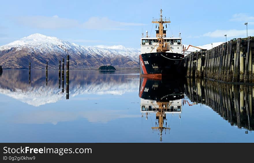 Ship in dock reflecting in clear blue waters with snow capped mountains in horizon with blue skies on sunny day.