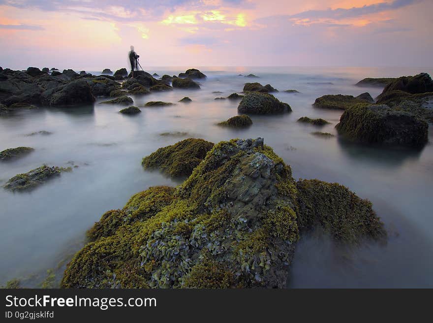 Blur of water over rocky shores with silhouette of standing person against sunset in cloudy skies. Blur of water over rocky shores with silhouette of standing person against sunset in cloudy skies.