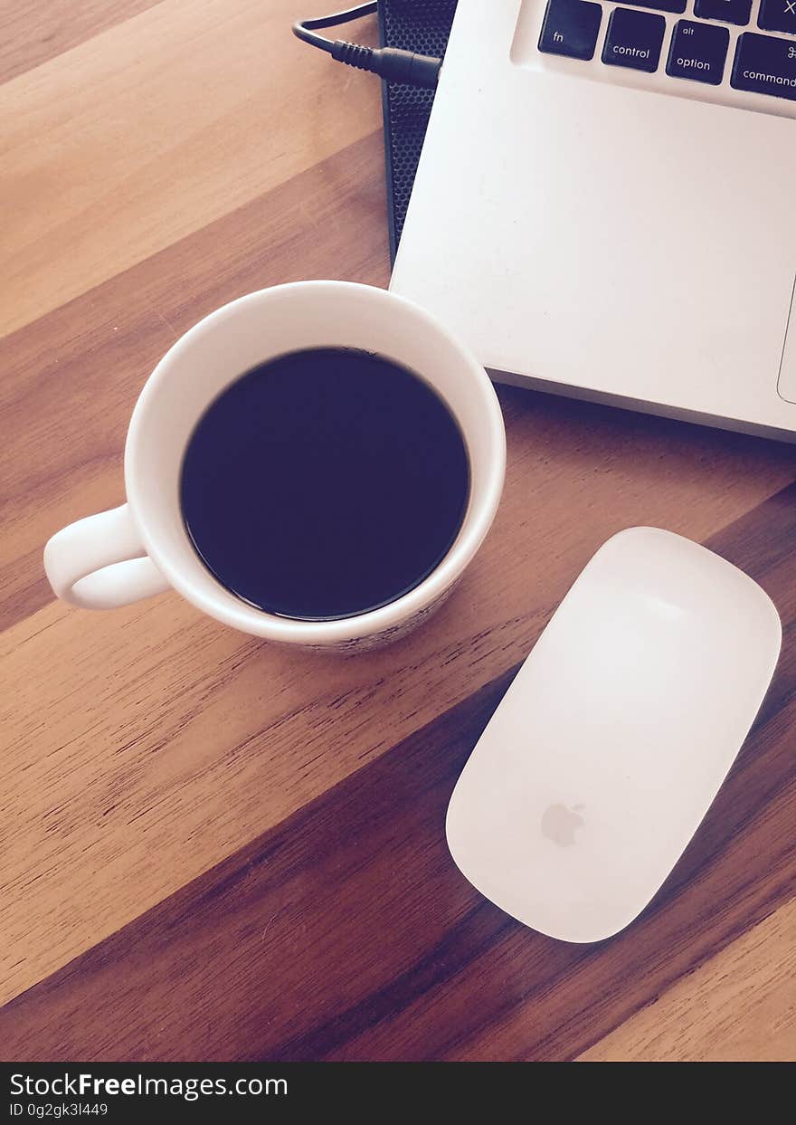 Overhead view of white china cup of coffee nest to laptop computer and Apple mouse on wooden tabletop. Overhead view of white china cup of coffee nest to laptop computer and Apple mouse on wooden tabletop.