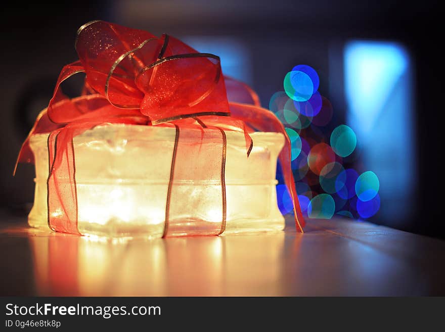 Clear glass gift box illuminated at night with red bow inside house with bokeh Christmas lights. Clear glass gift box illuminated at night with red bow inside house with bokeh Christmas lights.