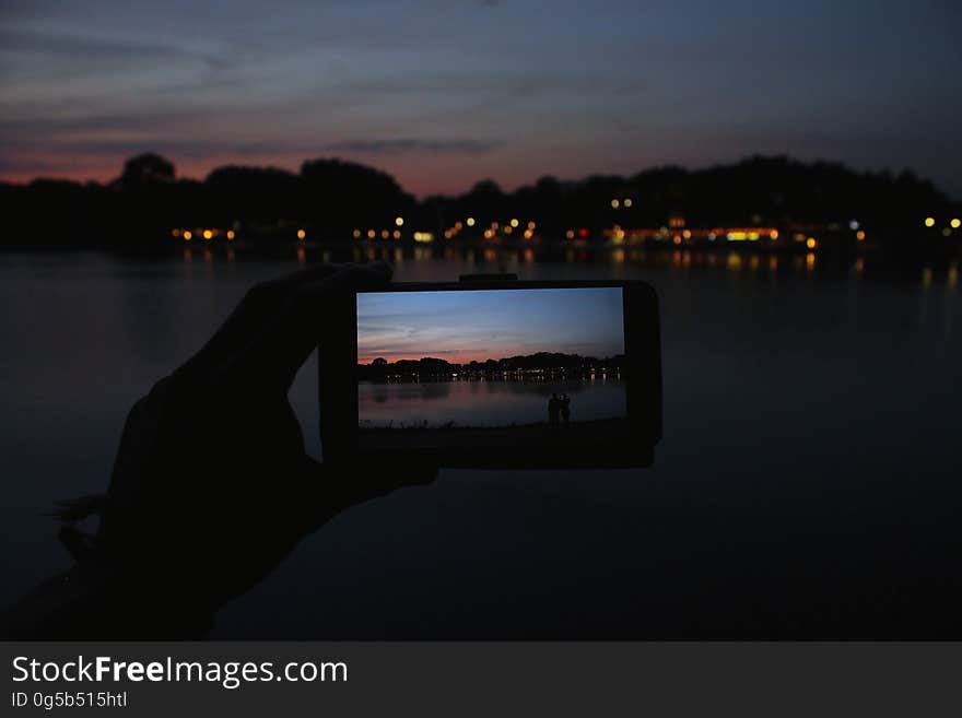 Hand holding smartphone with image of coastal sunset against landscape. Hand holding smartphone with image of coastal sunset against landscape.