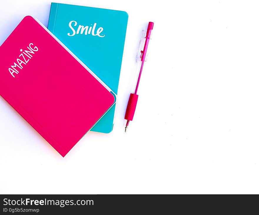 Two small notebooks, a red one marked "Amazing" and a blue one marked "Smile" alongside is a red pen, white background. Two small notebooks, a red one marked "Amazing" and a blue one marked "Smile" alongside is a red pen, white background.