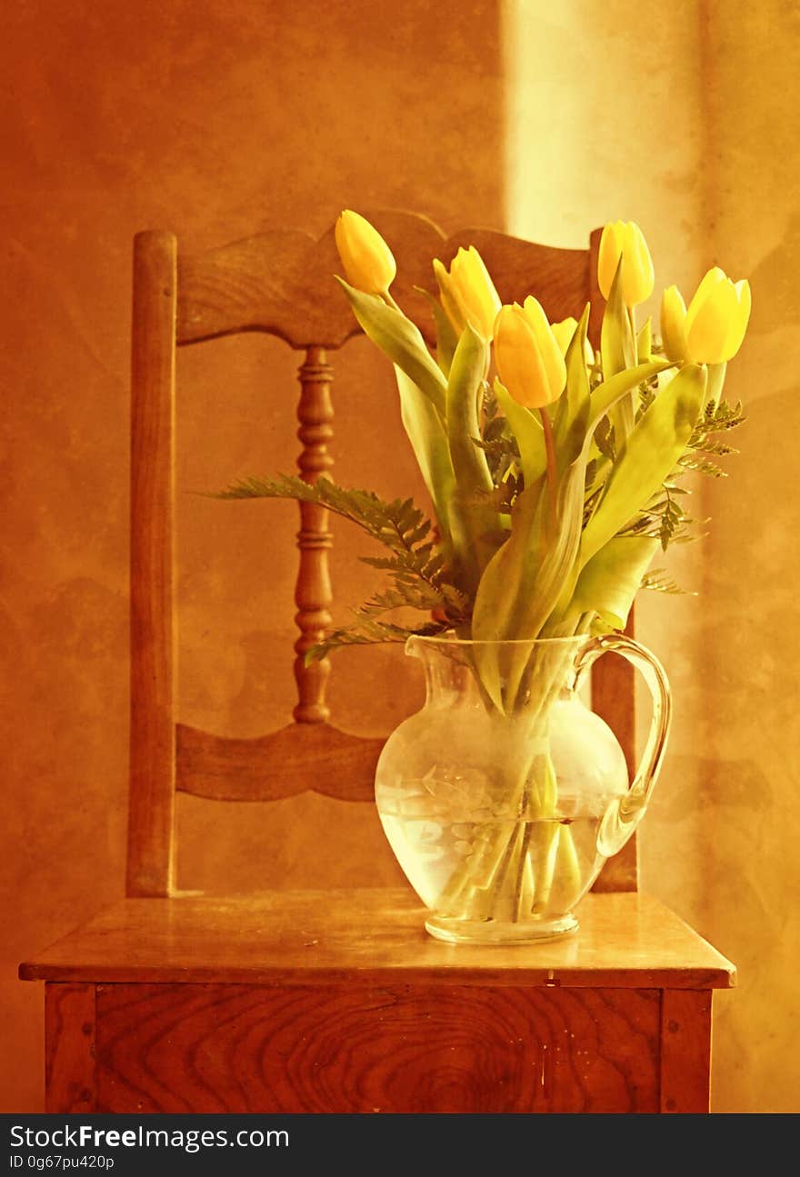 Glass pitcher vase with yellow tulips on small wooden table with chair in sunlight. Glass pitcher vase with yellow tulips on small wooden table with chair in sunlight.