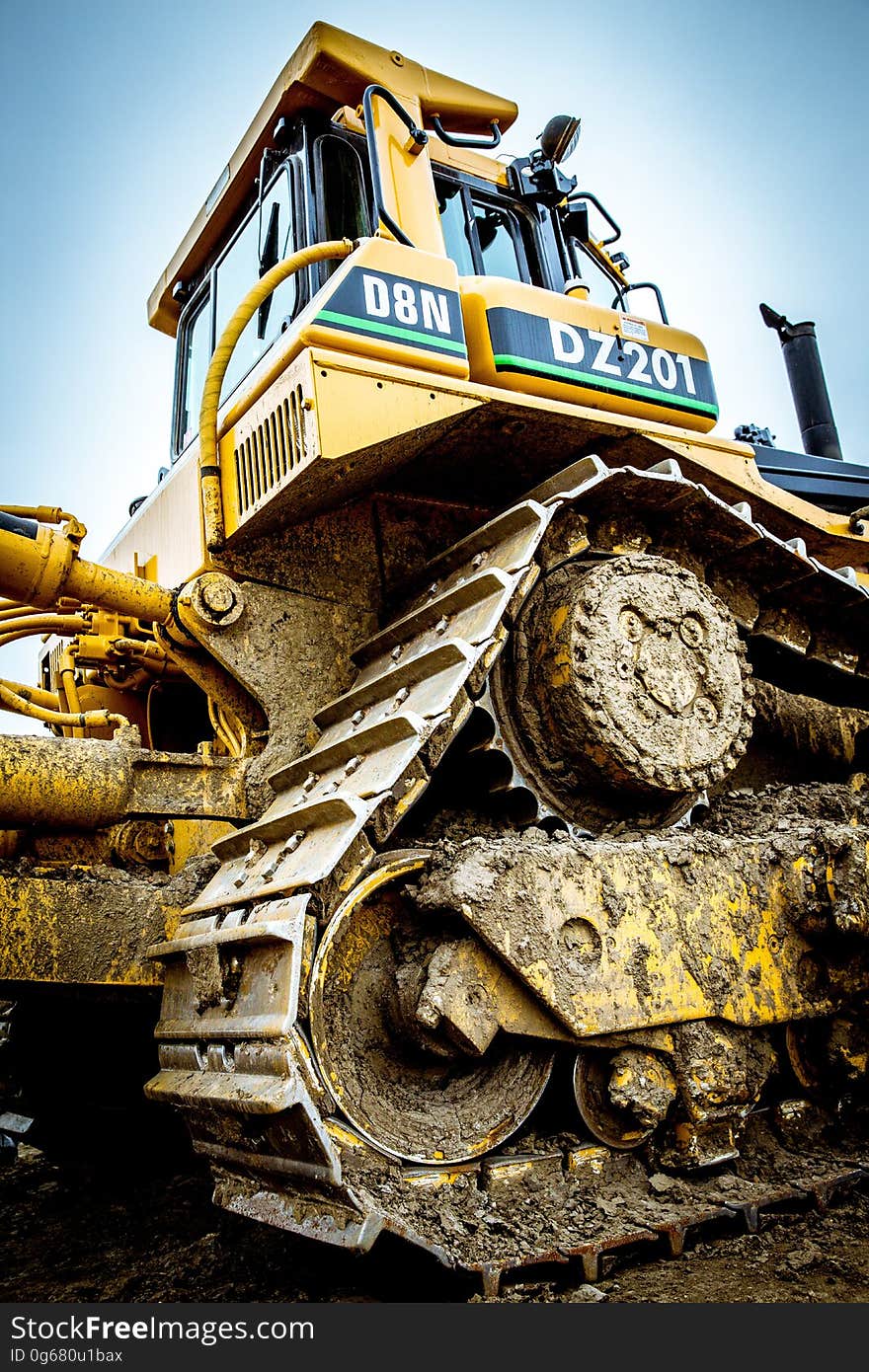 Closeup of bulldozer on construction site showing tracks and drive wheels (cogs) covered in mud and yellow weatherproof cab on top, pale blue sky. Closeup of bulldozer on construction site showing tracks and drive wheels (cogs) covered in mud and yellow weatherproof cab on top, pale blue sky.