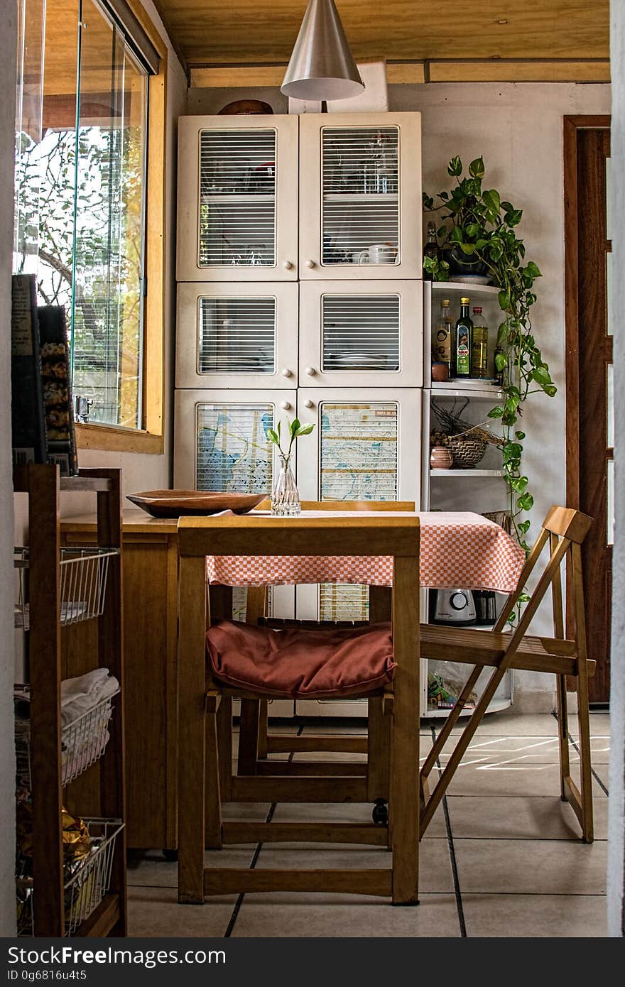 Dining table and chairs in the kitchen under a window looking out upon the garden with trees. White painted cupboards and shelves placed on the far wall supporting decorative plants. Dining table and chairs in the kitchen under a window looking out upon the garden with trees. White painted cupboards and shelves placed on the far wall supporting decorative plants.