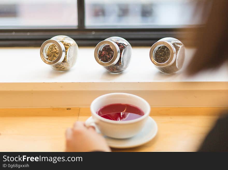 Hand of person holding cup of fruit tea with jars of herbs on ledge by window. Hand of person holding cup of fruit tea with jars of herbs on ledge by window.