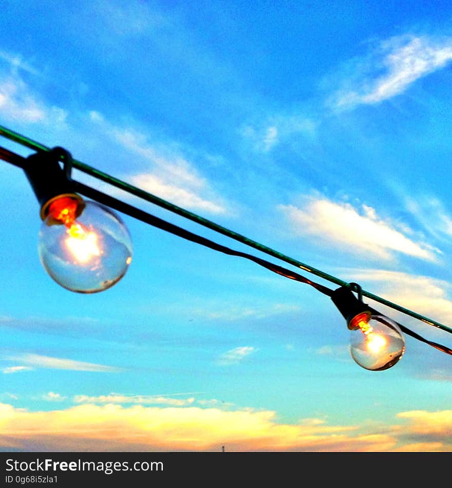 Two lighted bulbs hung outdoors at sunset with blue sky and cloudscape background. Two lighted bulbs hung outdoors at sunset with blue sky and cloudscape background.