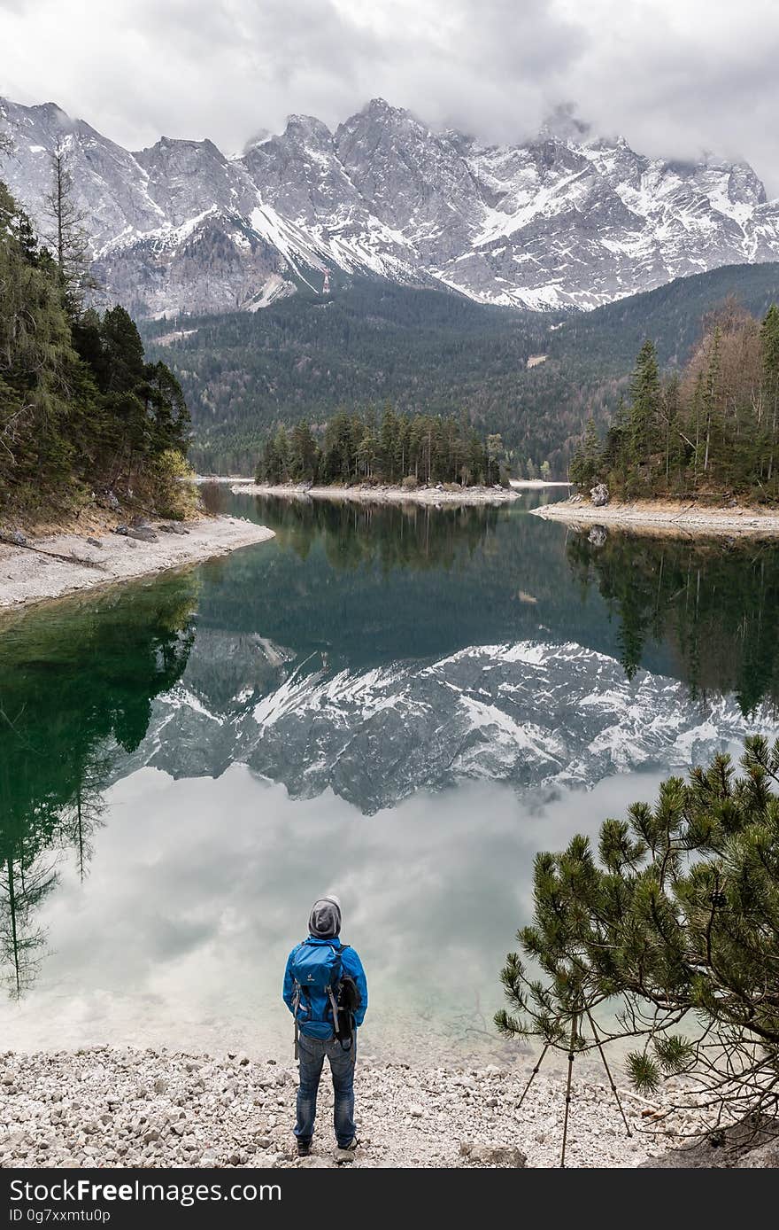 Hiker on banks of alpine lake with snow capped mountains reflecting in still waters.