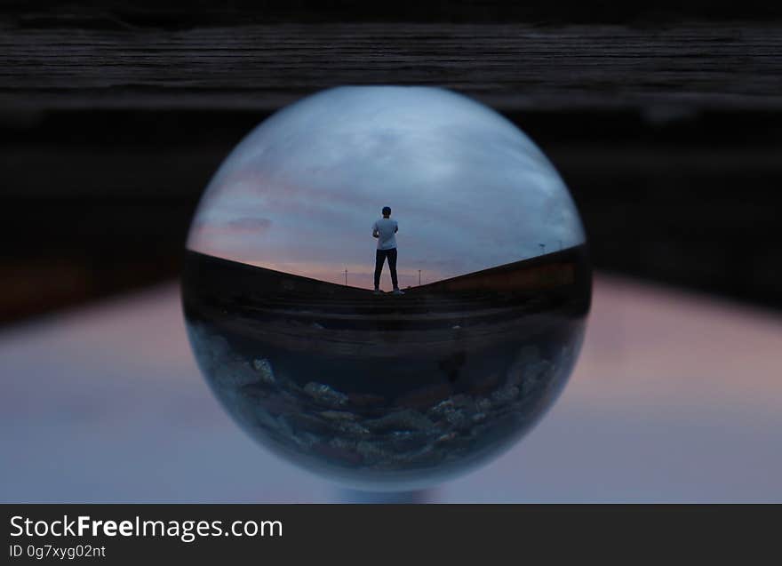 Glass sphere illuminated from above, and with the small reflected image of a man wearing shorts in it, upper background dark and lower part light gray. Glass sphere illuminated from above, and with the small reflected image of a man wearing shorts in it, upper background dark and lower part light gray.