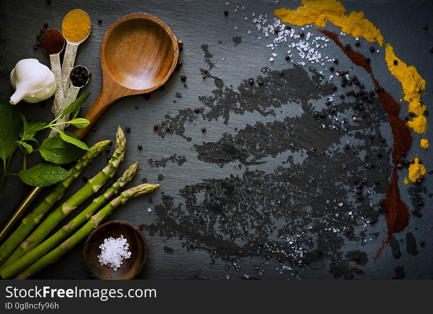 Food ingredients laid out on a dark surface including asparagus, garlic clove, mint leaves, herbs and paprika alongside spoons and a small bowl. Food ingredients laid out on a dark surface including asparagus, garlic clove, mint leaves, herbs and paprika alongside spoons and a small bowl.