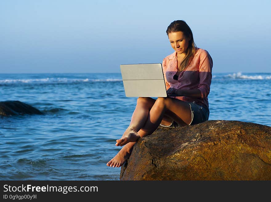 A woman working on a laptop while sitting on a rock at the seaside. A woman working on a laptop while sitting on a rock at the seaside.