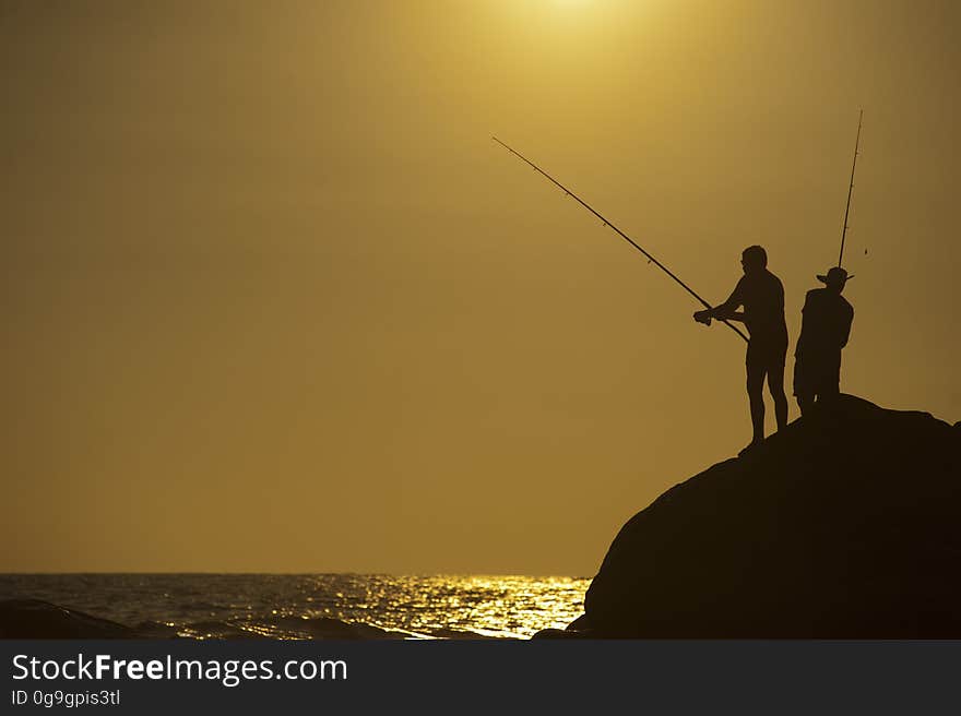 Two sea anglers using rod and line seen in silhouette standing on a large rock at dawn (or sunset), golden sky background. Two sea anglers using rod and line seen in silhouette standing on a large rock at dawn (or sunset), golden sky background.