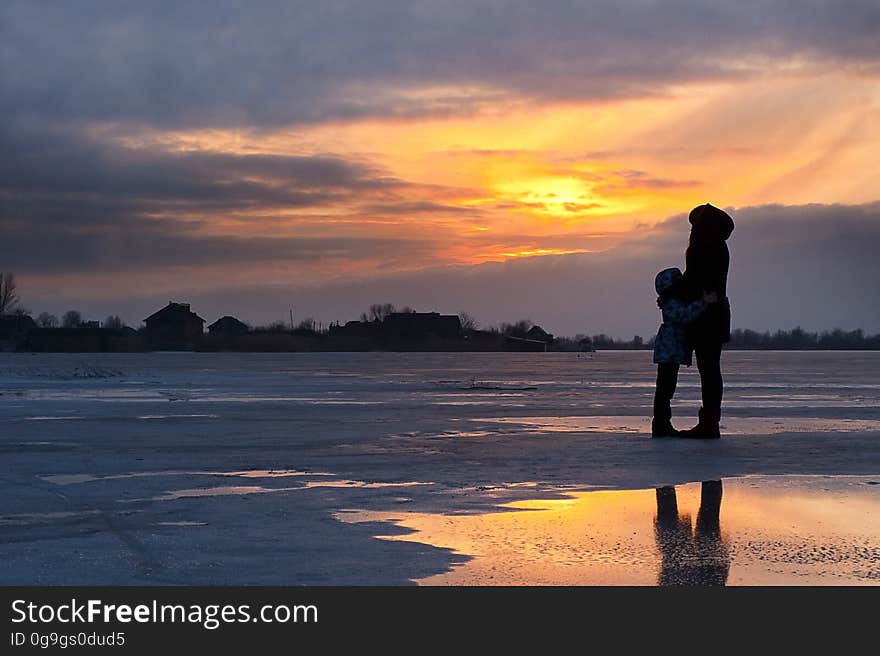 Silhouetted mother and child by frozen lake at sunset.