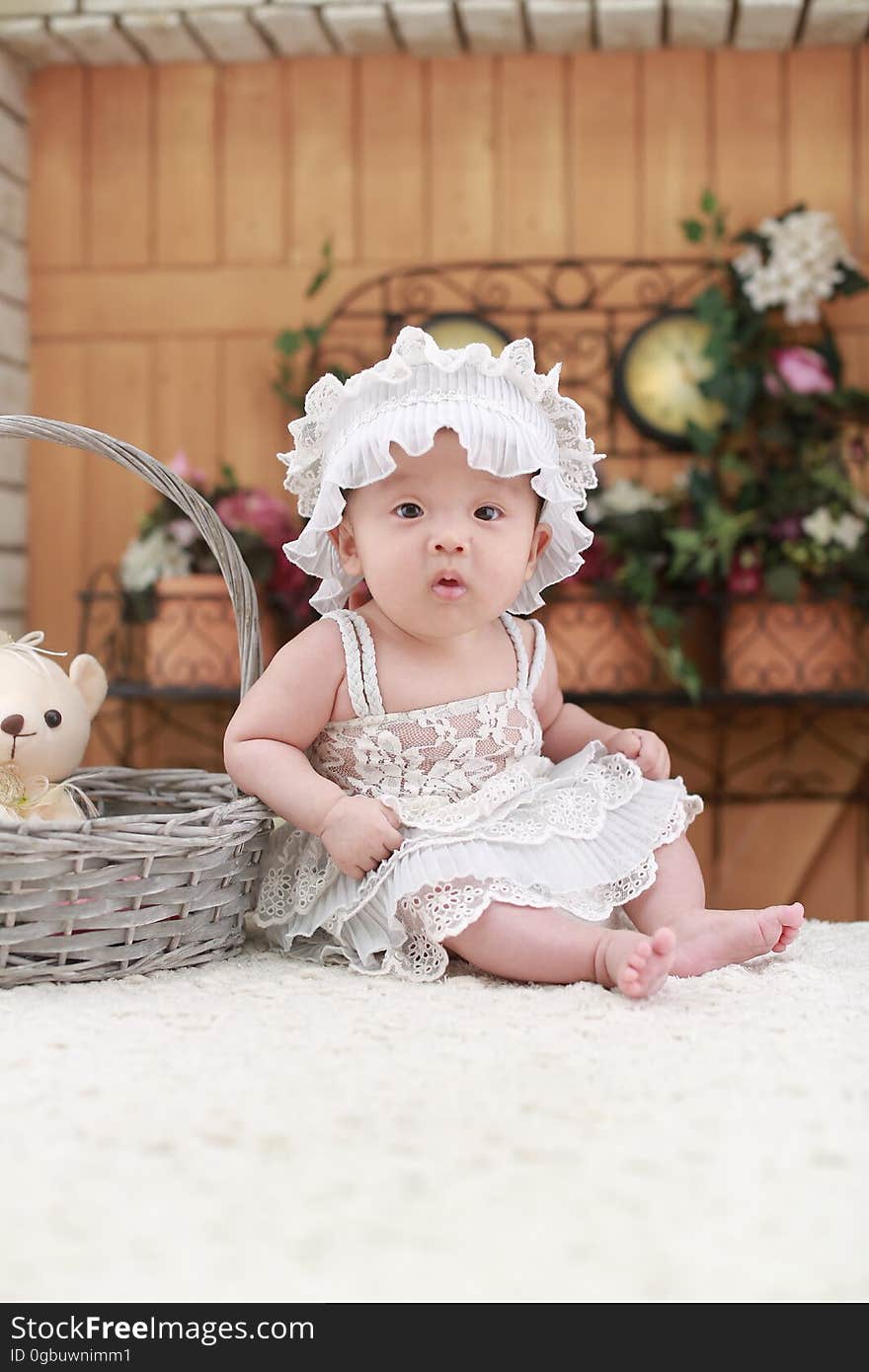 Baby Wearing White Headband and White Lace Floral Dress Sitting Beside Gray Wicker Basket