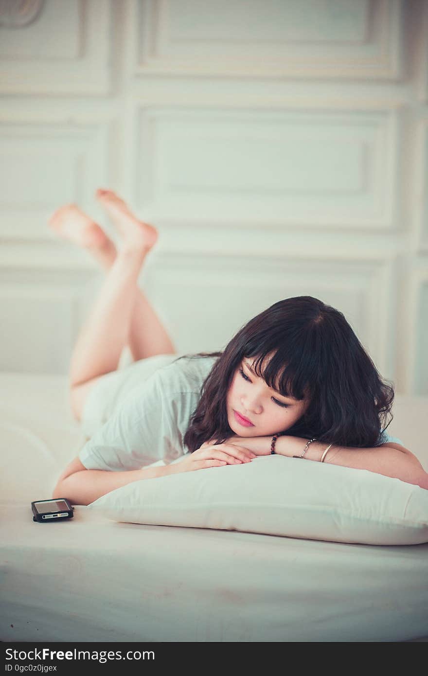 Pretty young Asian woman in a paneled room lying prone with her arms resting on a cushion, listening to music or chat from a black mobile phone. Pretty young Asian woman in a paneled room lying prone with her arms resting on a cushion, listening to music or chat from a black mobile phone.