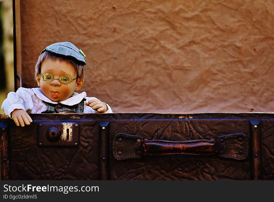 Antique baby boy (doll) with rosy cheeks dressed in white jacket and blue flat cap wearing spectacles standing behind a leather suitcase, mostly brown textured background. Antique baby boy (doll) with rosy cheeks dressed in white jacket and blue flat cap wearing spectacles standing behind a leather suitcase, mostly brown textured background.