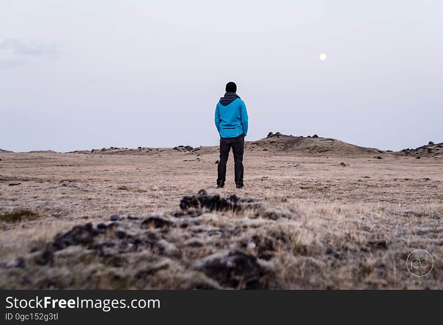 Rear portrait of man in blue jacket standing in bare field with moon in sky. Rear portrait of man in blue jacket standing in bare field with moon in sky.