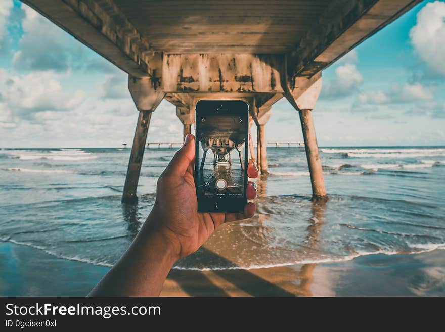 Hand holding mobile phone to take a picture while the person is in the sea under a wooden jetty on legs, background pale blue sky and clouds. Hand holding mobile phone to take a picture while the person is in the sea under a wooden jetty on legs, background pale blue sky and clouds.