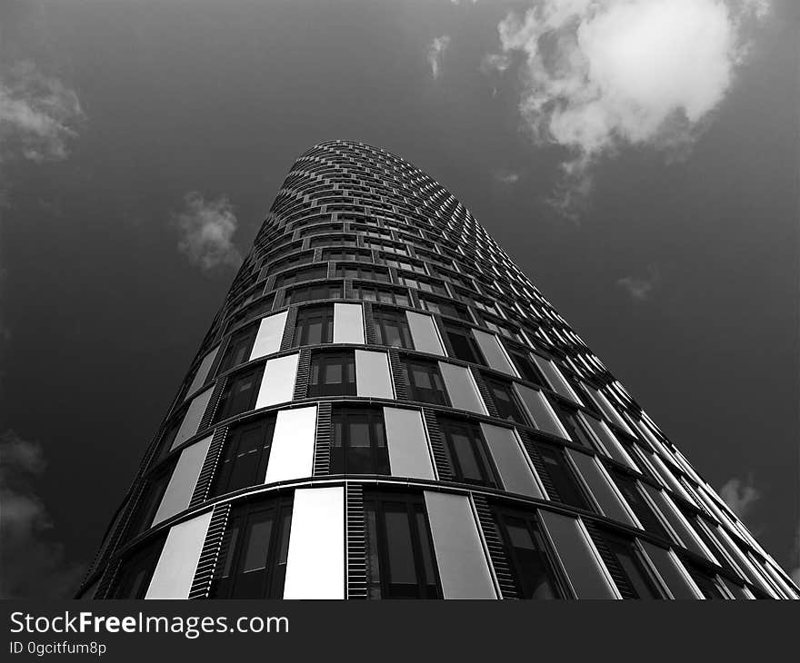 Unusual building, skyscraper built of concrete, steel and glass but with curved facade and seen from (above) ground pointing upwards, gray sky and cloud. Unusual building, skyscraper built of concrete, steel and glass but with curved facade and seen from (above) ground pointing upwards, gray sky and cloud.