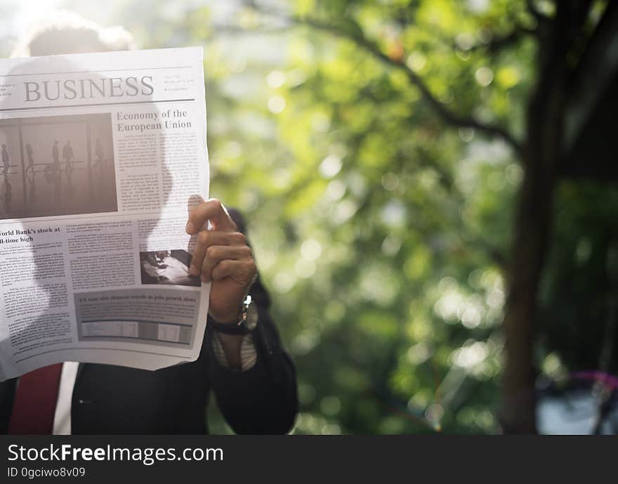 Man in a business suit reading the business section of a newspaper outdoors by trees, with his face obscured and his silhouette visible on the newspaper from the sunlight behind. Man in a business suit reading the business section of a newspaper outdoors by trees, with his face obscured and his silhouette visible on the newspaper from the sunlight behind.