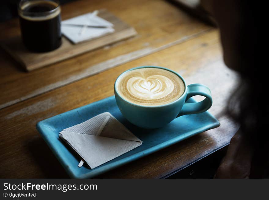 wooden table with cup of coffee in stylish matching blue rectangular shaped saucer with heart shape in foam on the top and napkin square in the saucer. wooden table with cup of coffee in stylish matching blue rectangular shaped saucer with heart shape in foam on the top and napkin square in the saucer.