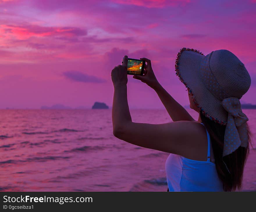 Rear view of a young woman making picture on cell telephone camera while enjoying her vacation holidays on the seashore at sunset, hipster girl photographing amazing sea landscape on her smart phone